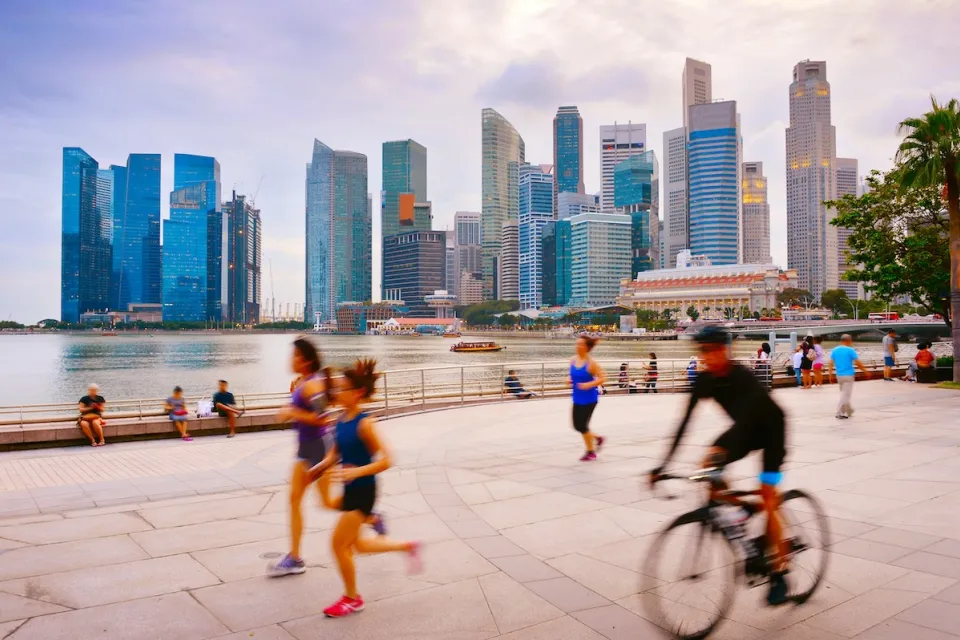 People running and cycling on wide paved path next to water with Singapore skyline with high-rises in background.
